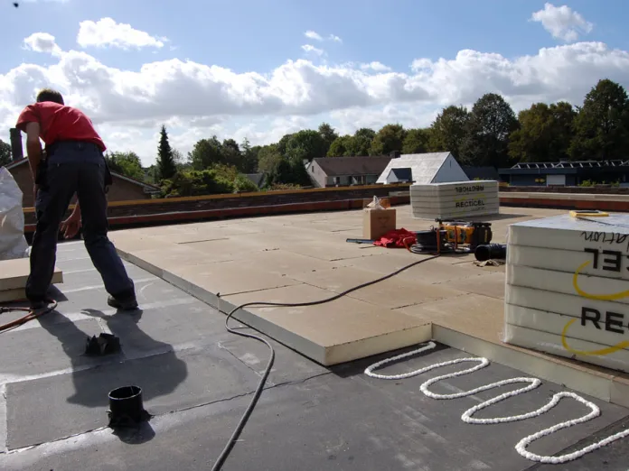 Worker on flat roof with construction materials, under partly cloudy sky.