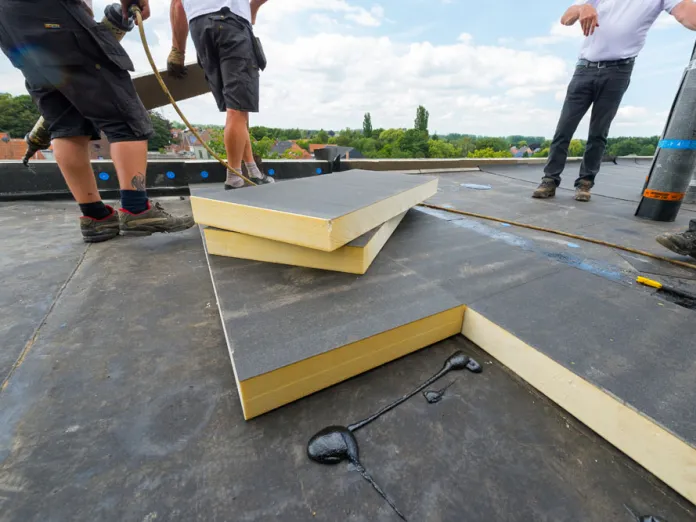 Workers installing foam insulation on a flat rooftop under a blue sky.