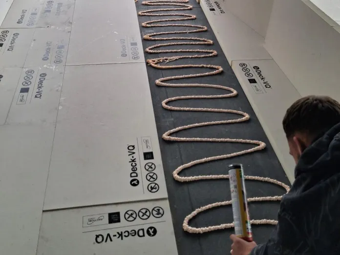 Person applying adhesive in zigzag pattern on a decking sheet.