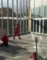 Workers in safety gear fixing flooring in a large, sunlit room with glass walls.