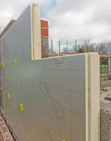 Building wall with insulation panels and brick foundation under a cloudy sky.