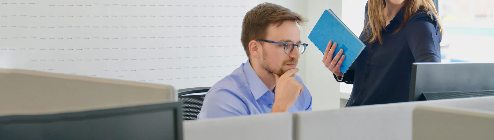 A man at a desk and a woman standing, both looking at a computer screen.