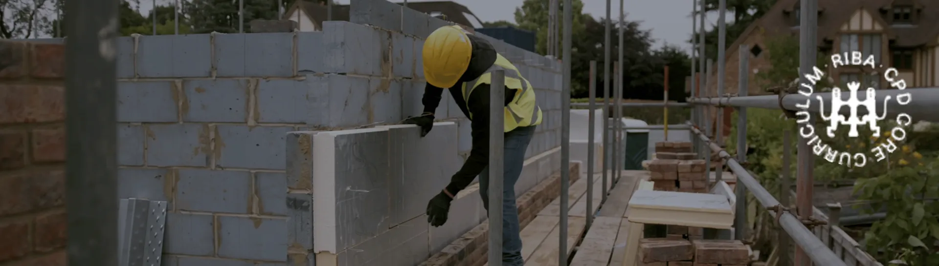 Builder laying bricks on scaffolding, wearing a yellow helmet and safety vest.
