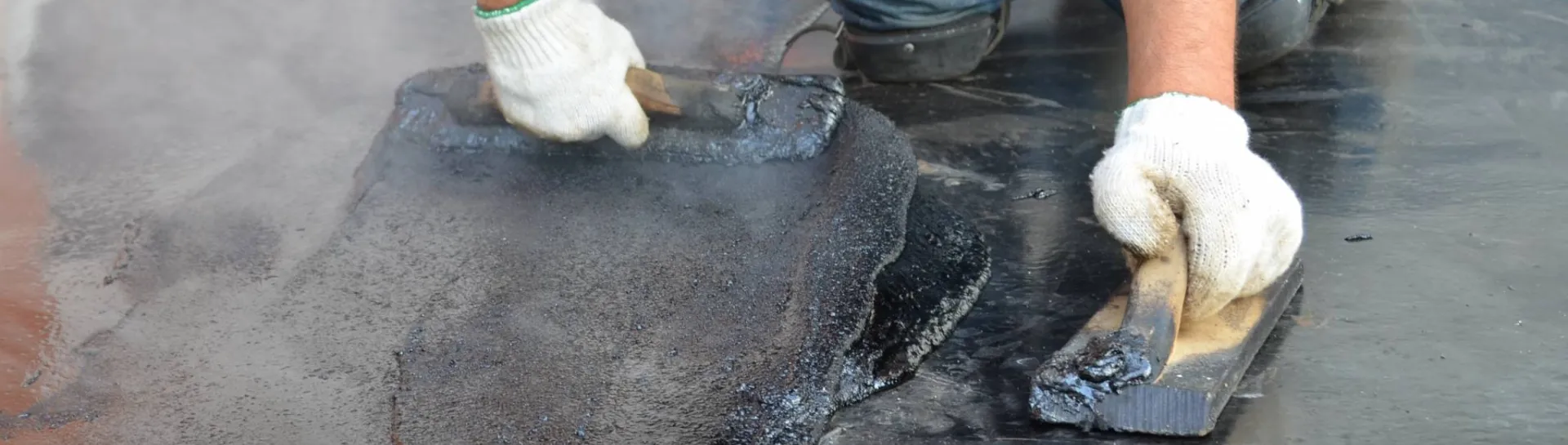 Worker applying hot tar with metal tools on a flat roof.