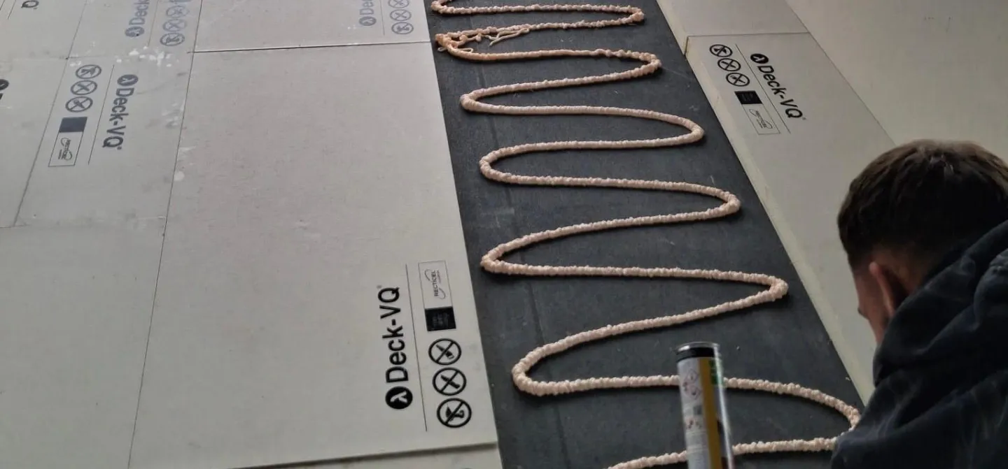 Person applying adhesive in zigzag pattern on a decking sheet.