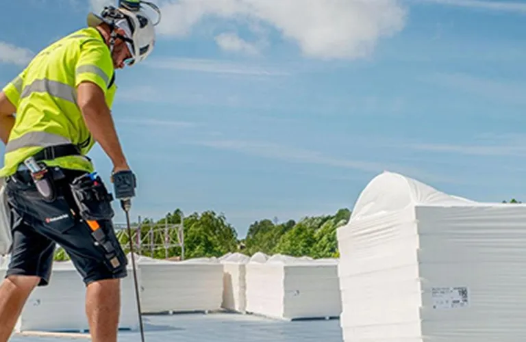 Bouwvakker in veiligheidskleding werkt op plat dak onder blauwe lucht.