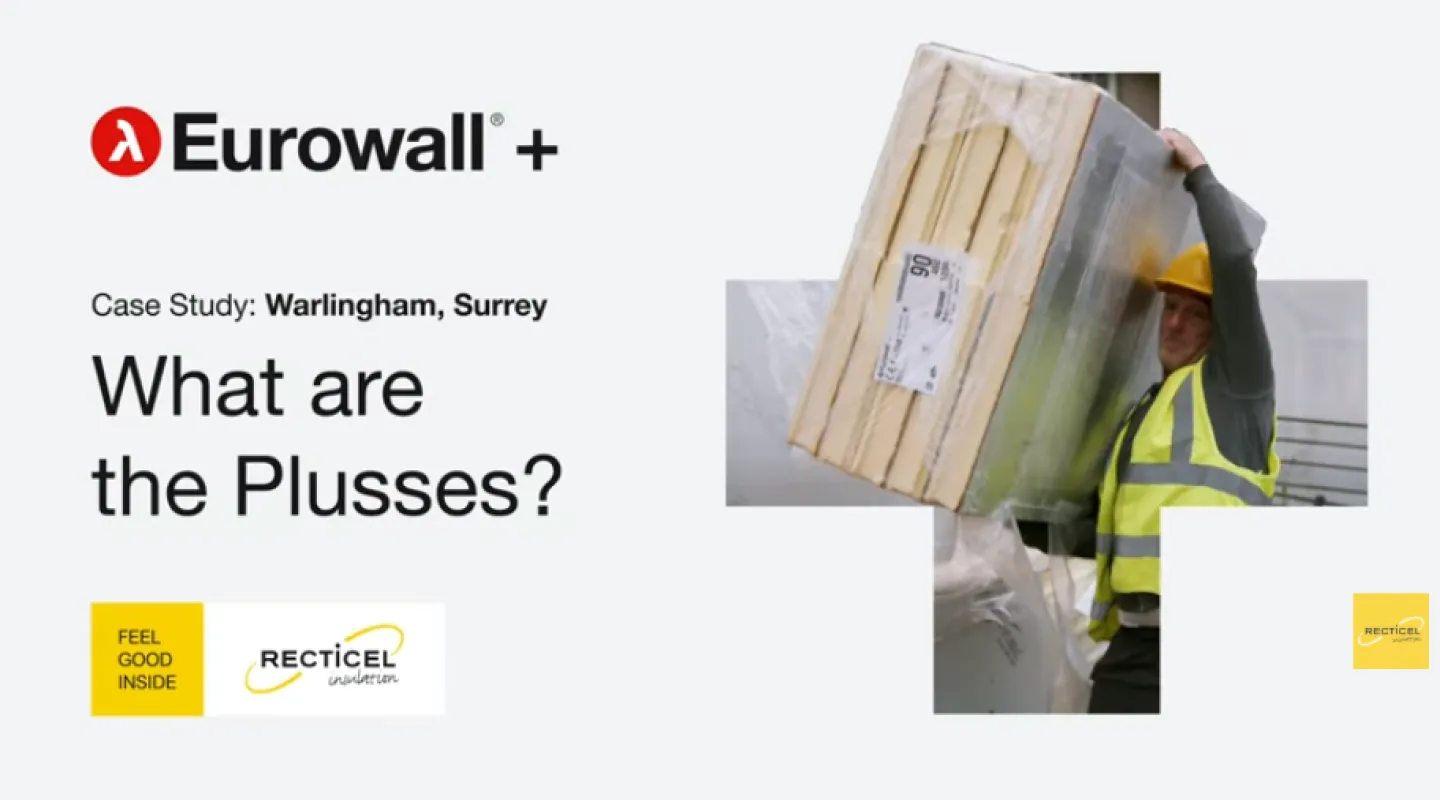 Construction worker carries insulation panels, wearing a hard hat and hi-vis vest.