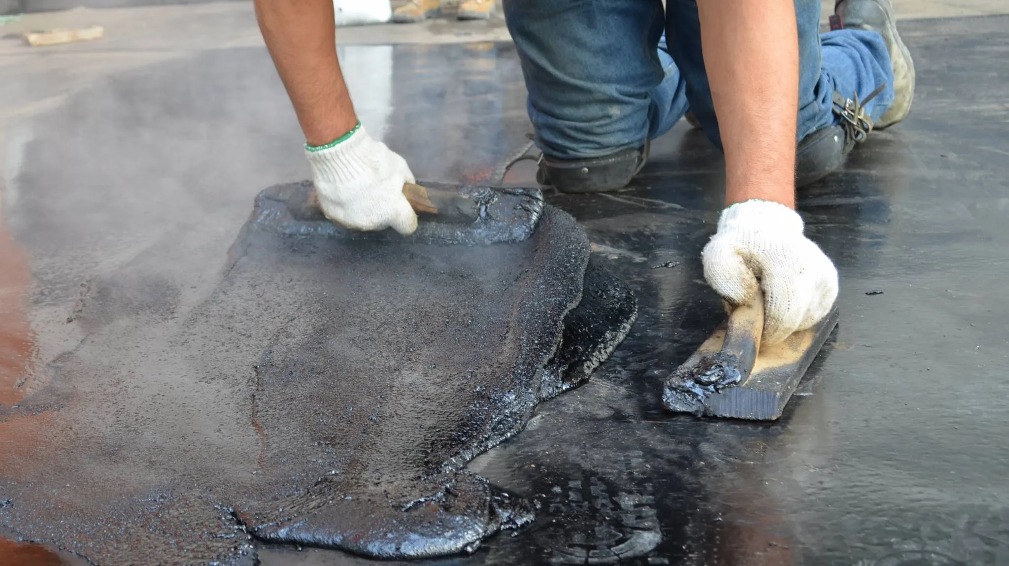 Worker applying hot tar with metal tools on a flat roof.