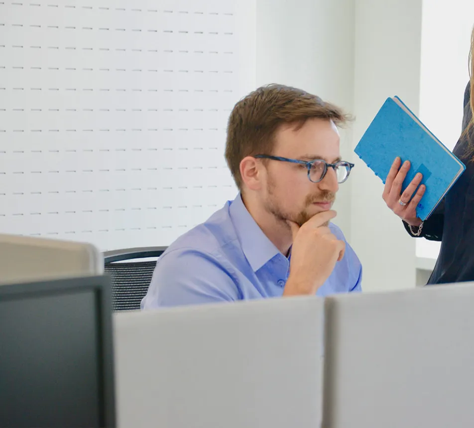 A man at a desk and a woman standing, both looking at a computer screen.