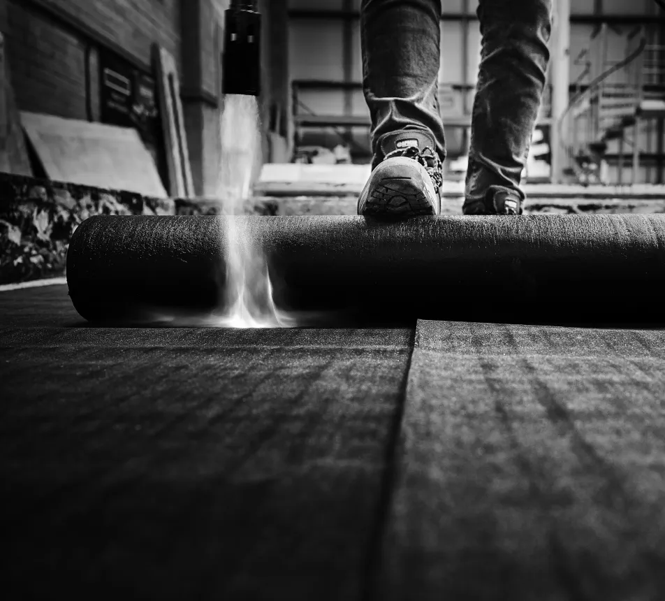 Black and white photo of a person rolling out roofing material indoors.