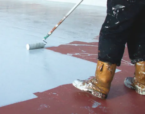 Person painting floor grey with roller, wearing boots and black trousers.