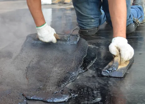 Worker applying hot tar with metal tools on a flat roof.